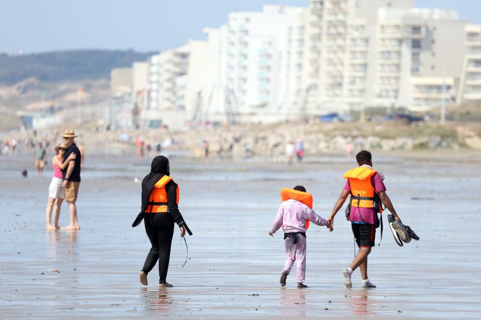 Migrant families, including children, in orange life jackets walking on a beach.