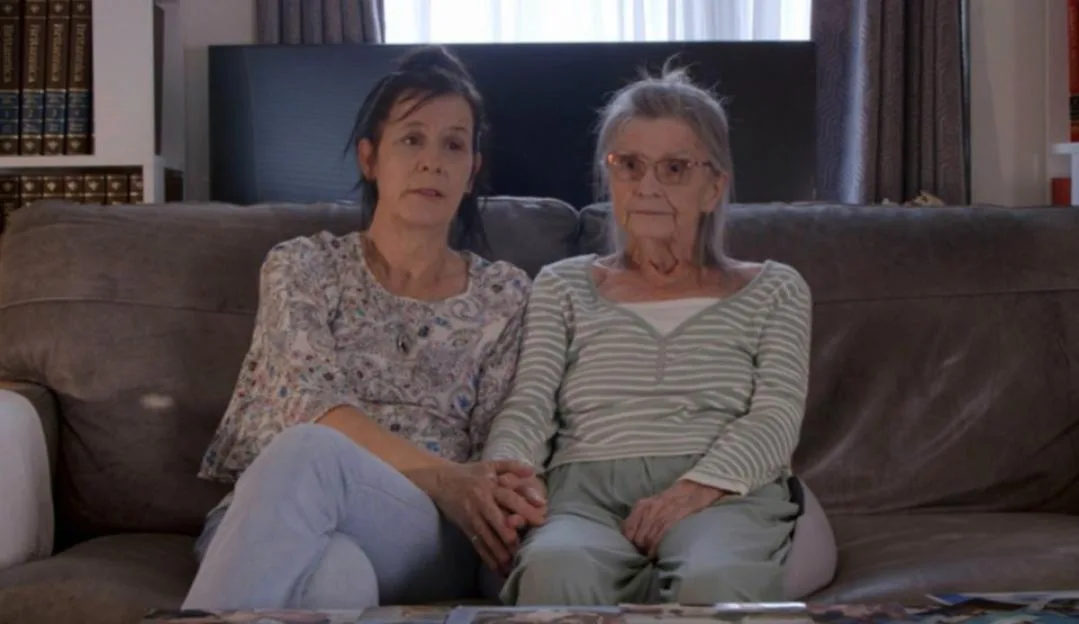 Two women, one younger and one older, sit on a couch, holding hands.