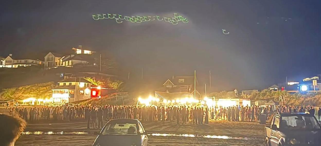 Crowd of people gathered on Polzeath beach at night, with two cars in the foreground and distant houses on a hill.