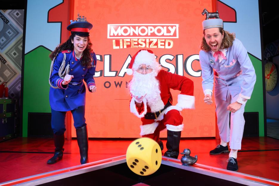 Three actors portraying Monopoly characters, including Santa Claus, on a stage with a giant die and duck token.