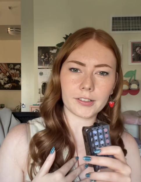 A woman with red hair, green nail polish, and a strawberry earring holds a device that appears to be a multi-color light.