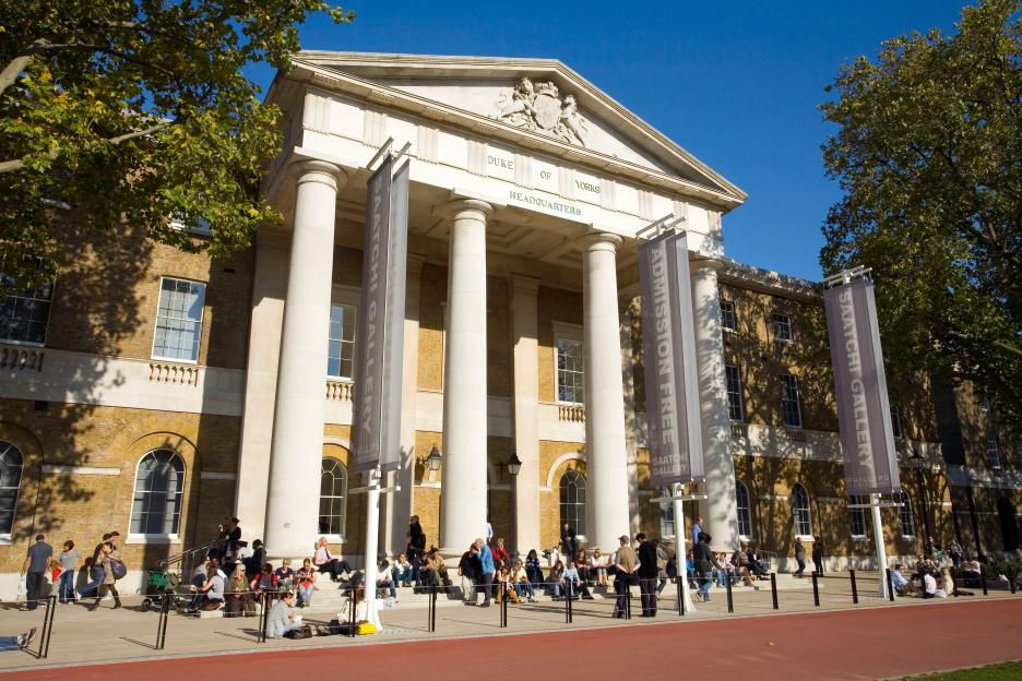 The Saatchi Gallery in London, with "ADMISSION FREE" banners displayed, on a sunny day with people sitting on the steps outside.