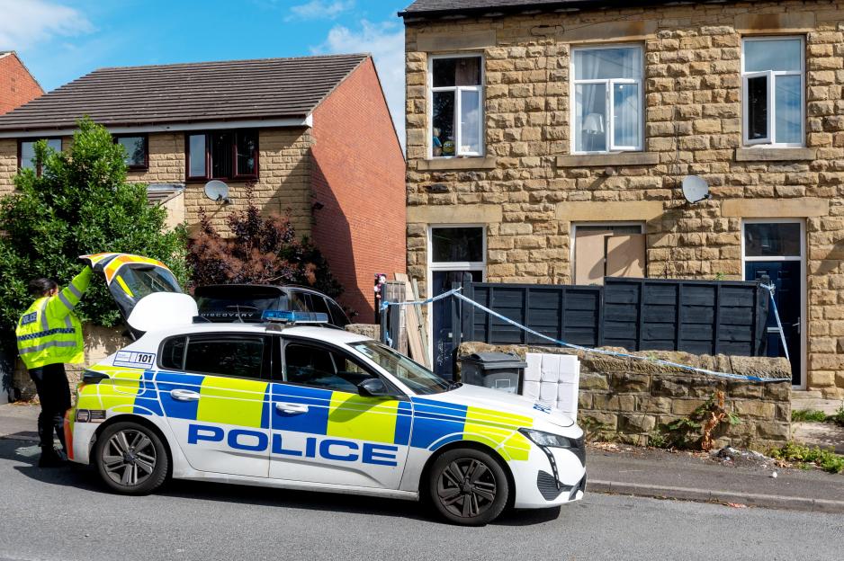 A police officer stands by an open police car trunk in front of a stone house with blue and white police tape across the entrance.