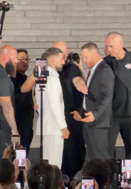 Two men, one in white and one in black, facing each other with several people and phones around them.