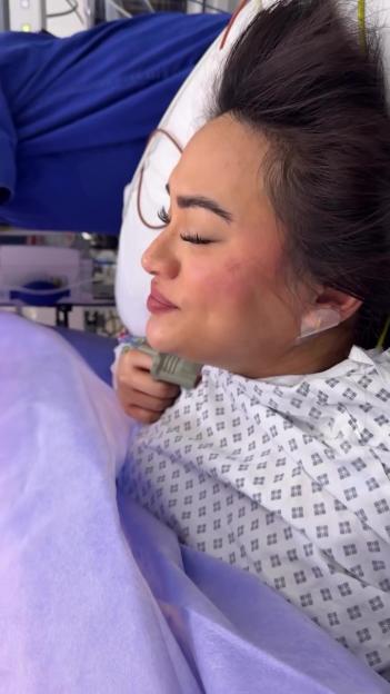 A woman in a hospital gown and makeup lying in a bed with her eyes closed.