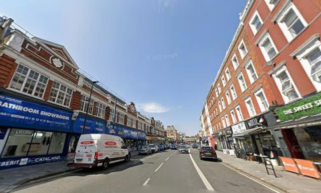 Street lined with shops and apartments, with a white van parked in front of a "Bathroom Showroom" and cars driving on the road.