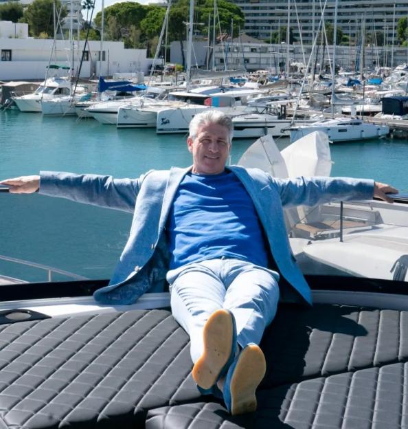 Man relaxing on a boat with arms outstretched, overlooking a marina with yachts and buildings in the background.
