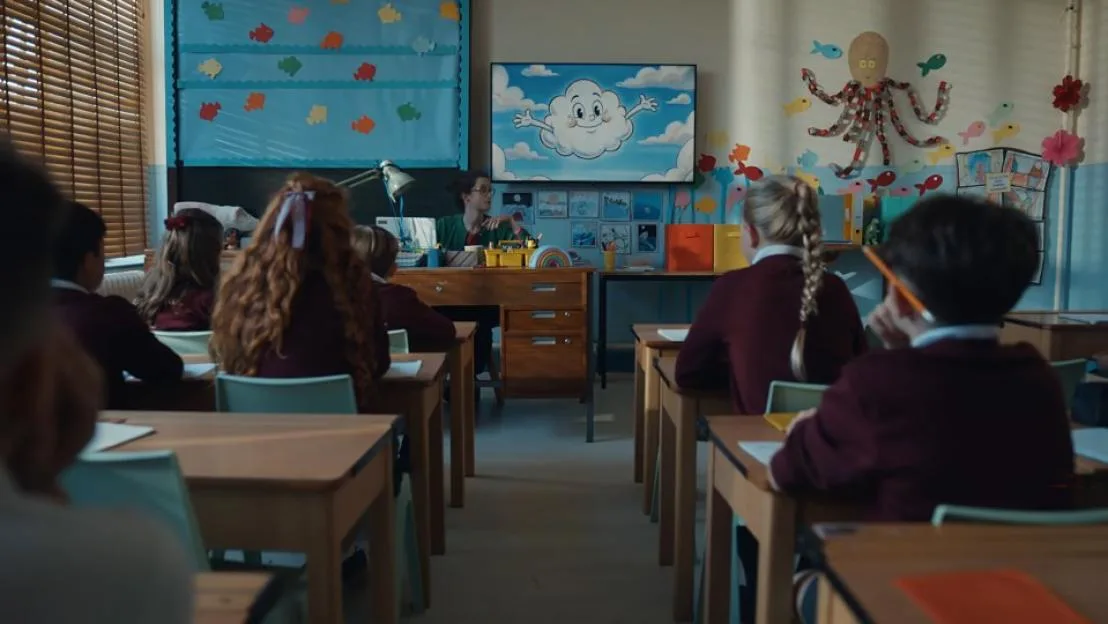 Students in a classroom listen to a teacher while a screen in the front displays a cartoon cloud.