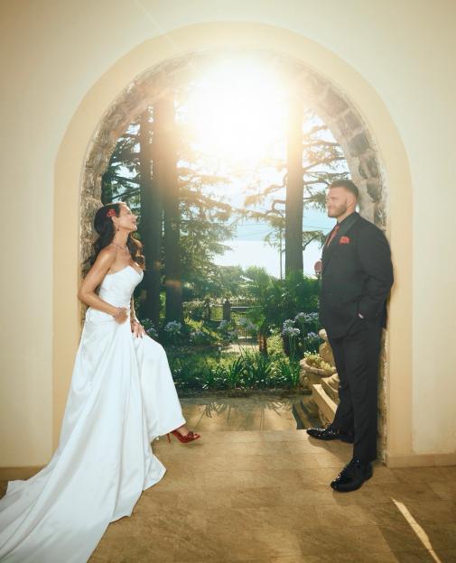 A bride in a white gown and red shoes, and a groom in a black suit with a red pocket square, posing in an arched doorway overlooking a sunny garden.