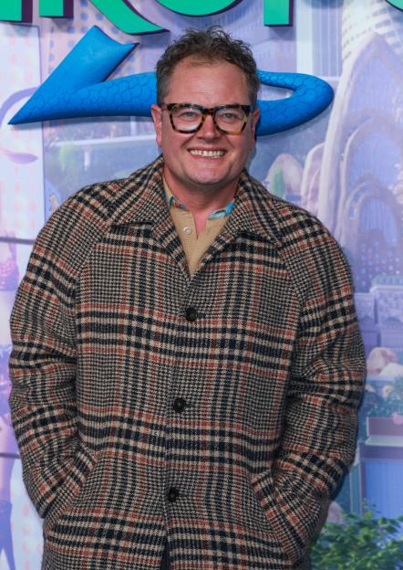 Alan Carr smiles in a brown plaid coat at the Zootropolis II premiere.