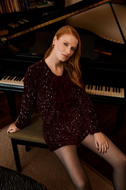 A woman in a sequined dress with a velvet bow posing by a grand piano.