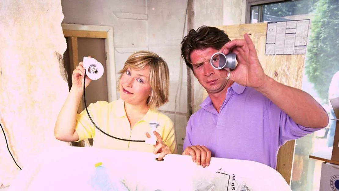 A woman and a man holding plumbing parts in a room undergoing renovation.