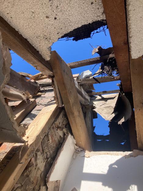 A large hole in a roof, showing damaged wooden beams, debris, and blue sky.