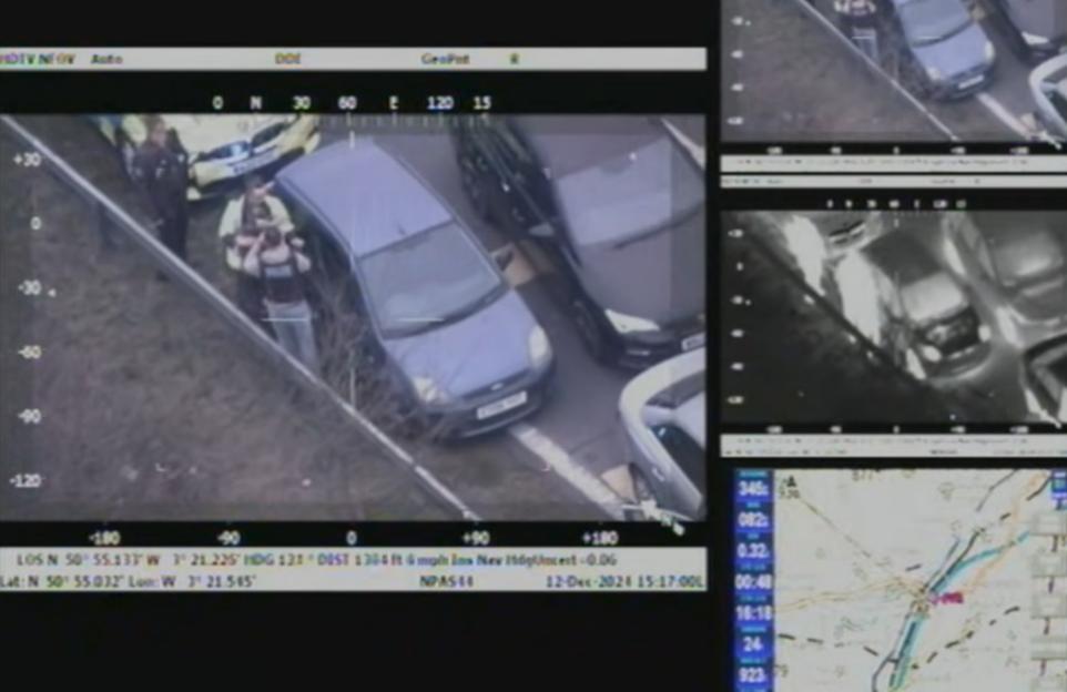 An aerial view of the arrest of Neil Warren, showing a police car, two police officers, and a civilian next to a parked blue car.