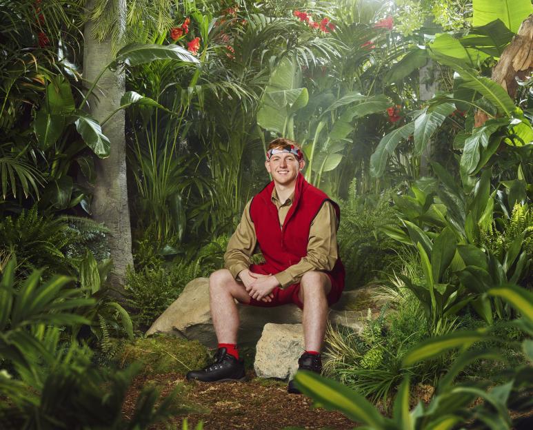 Angry Ginge in a red vest and shorts, sitting on a rock in a jungle setting.