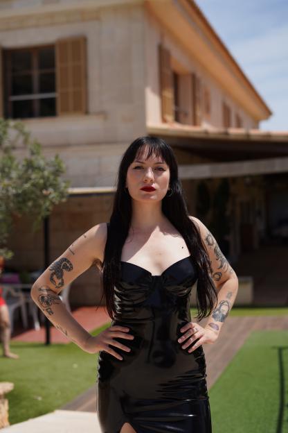 Paulita Pappel, founder of Lustery, in a black latex dress, with tattoos on both arms, standing in front of a house.