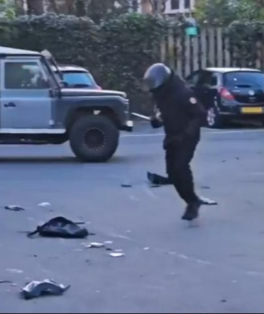A man in a helmet and dark clothing holds a machete, with debris on the ground, and a Land Rover in the background.