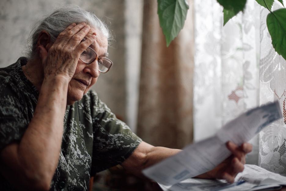 A worried senior woman with gray hair and glasses, holding a hand to her forehead while looking at bills.