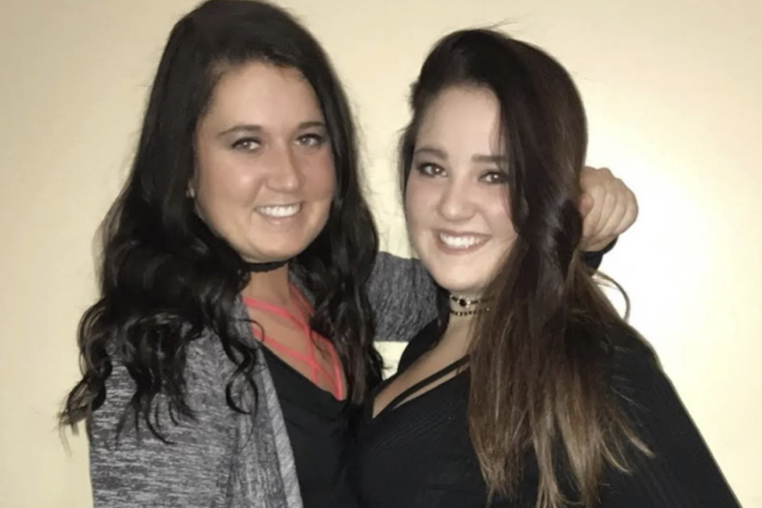 Two young women with long dark hair and chokers smiling at the camera, one with her arm around the other.