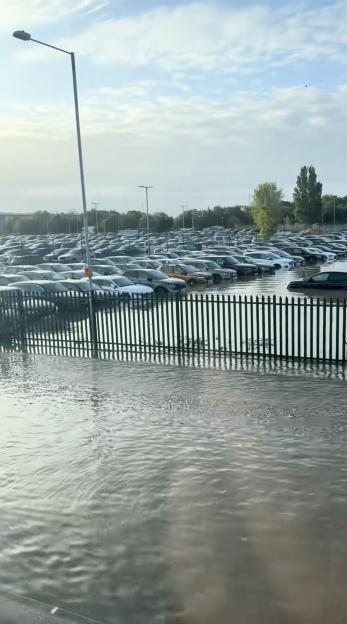 Video still of a flooded car park at Heathrow Airport, with numerous cars partially submerged in water behind a metal fence.