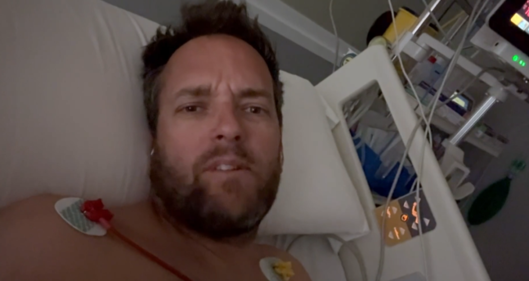 A man with a beard lying in a hospital bed with medical sensors on his chest.
