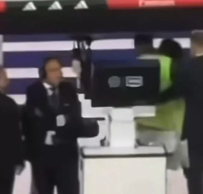 A man in a suit looks toward a VAR monitor while people argue in the background.