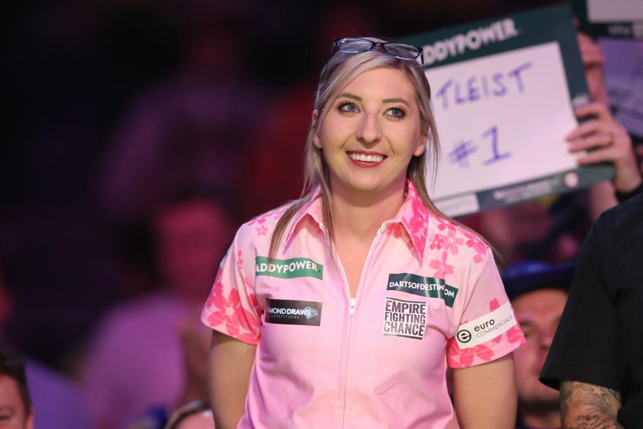 Fallon Sherrock smiling and looking up, wearing a pink shirt with cherry blossoms and sponsor logos.