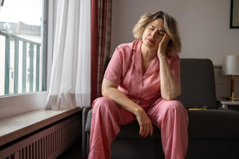 Mature woman sitting on a couch in pink pajamas, holding her head with her hand while suffering from a headache.