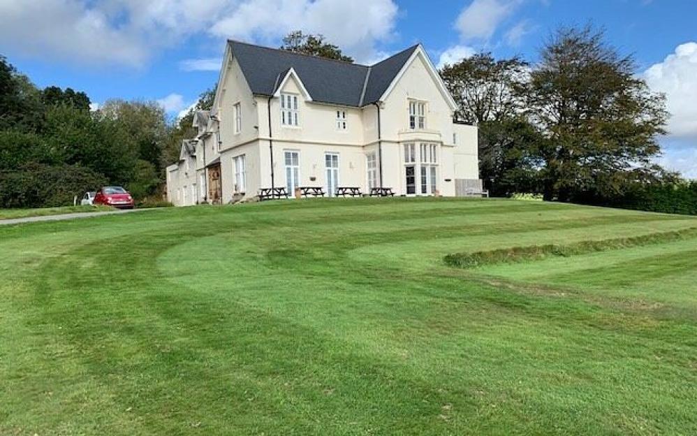 Welbeck Manor in Sparkwell, a large cream-colored building with multiple windows and a dark roof, surrounded by green lawns and trees.