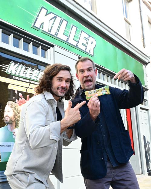 Joe Wicks and another man holding a "Killer" protein bar, pointing to it, in front of a store with a large "Killer" sign.
