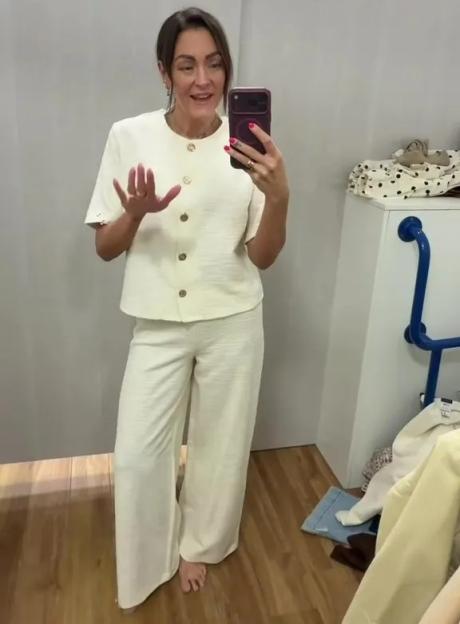 A woman in a cream-colored short-sleeved top and matching wide-leg pants is taking a mirror selfie.