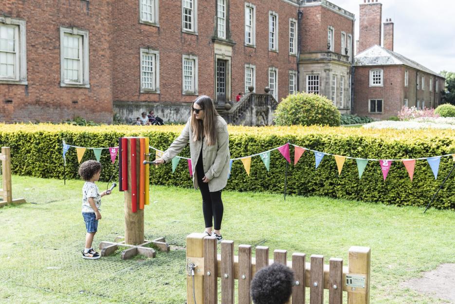 A woman and child playing with outdoor musical instruments in a garden with a large brick building in the background.