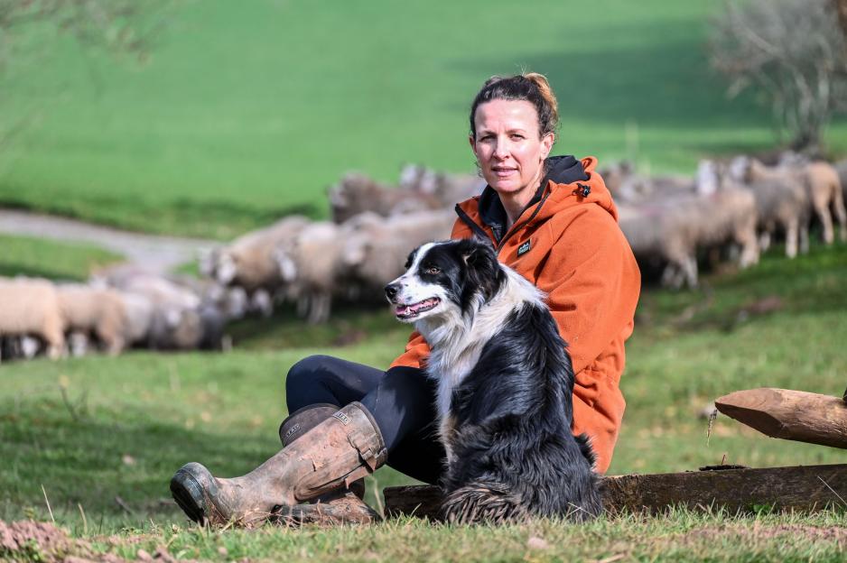 Bryony Gittins, a farmer, sits with her Border Collie as sheep graze in the background.