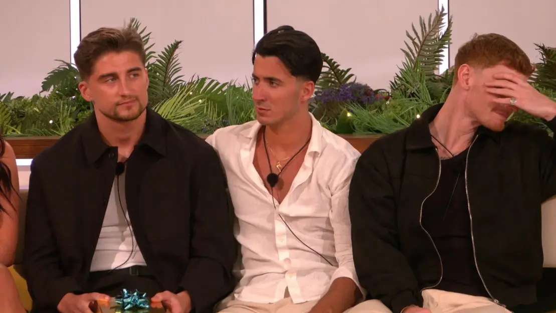 Three men sitting on a couch, one holding a gift box, another looking to his right, and the third covering his face with his hand.