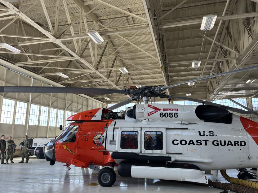 A U.S. Coast Guard helicopter, painted orange and white, sits in a hangar with military personnel standing nearby.