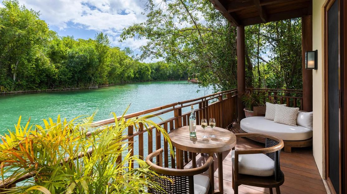 Fairmont Mayakoba balcony overlooking a turquoise river surrounded by lush green trees, with a table, chairs, and daybed.