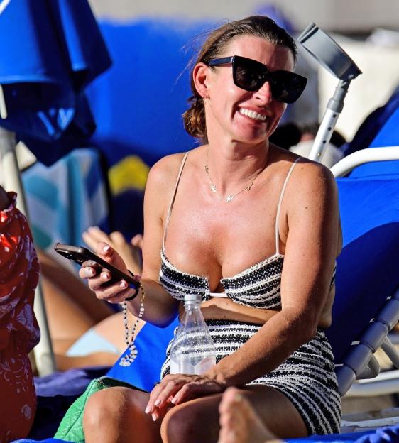 Coleen Rooney sitting on a beach chair in a black and white bikini, smiling and holding a phone.