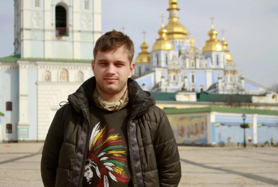 Bohdan Yermokhin, a young man who was deported by Russian forces, stands in front of a blue and gold domed building in Kyiv, Ukraine.