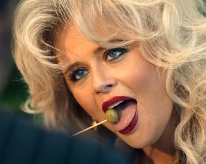 A woman with blonde hair, red lipstick, and blue eyes licking an olive on a toothpick.