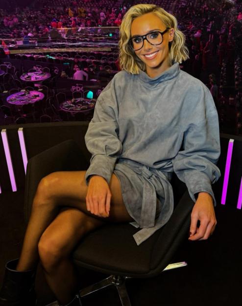 A woman with short blonde hair and glasses smiles while sitting in a chair.