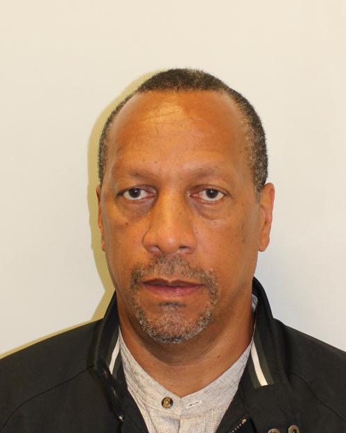 A headshot of David Checkley, a man with dark skin, wearing a dark jacket over a light gray collared shirt. He has short, dark, receding hair, a short graying goatee, and a serious expression.