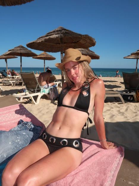 A woman in a black bikini and straw hat sits on a pink towel on a sandy beach.