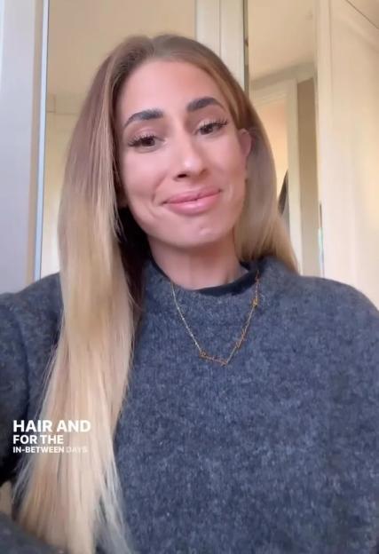 A woman with long blonde hair and a gray sweater smiles while wearing a gold necklace.