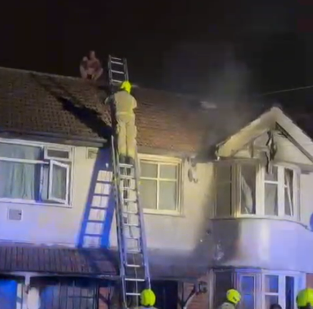 Firefighters using ladders to rescue a person from the roof of a burning house at night.