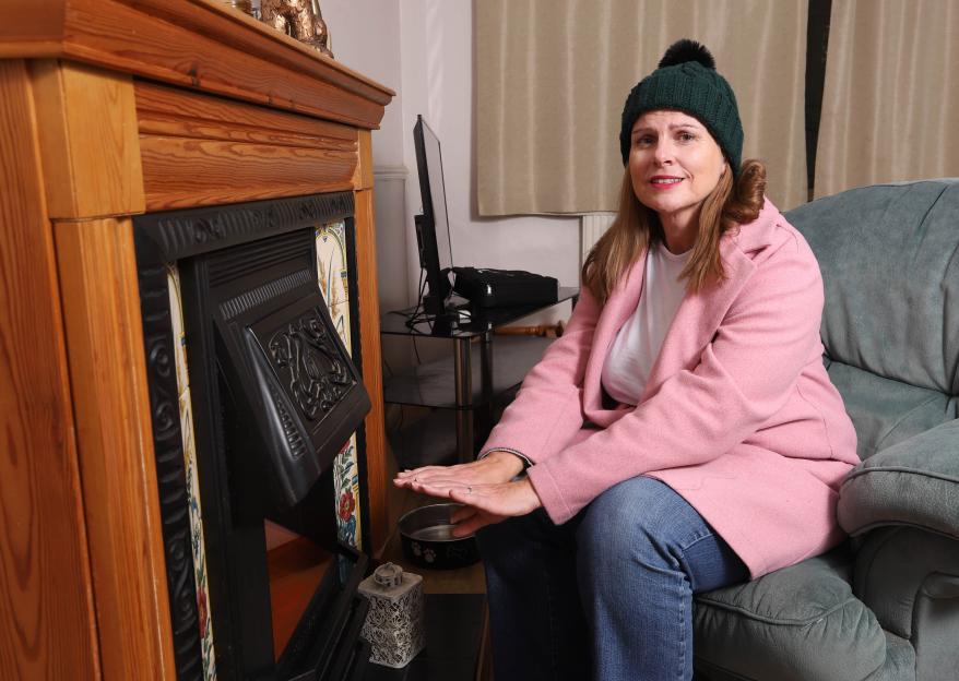 Ashley Peace, dressed in winter attire, warms her hands by an indoor fireplace.