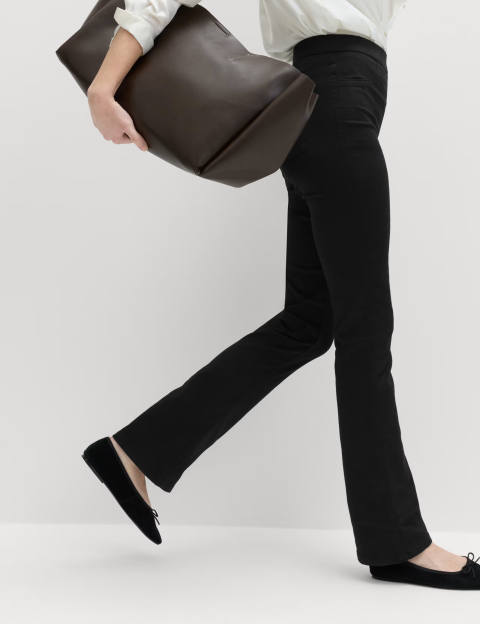 A person wearing black pants, black flats, and a white shirt holds a brown tote bag and walks to the right.