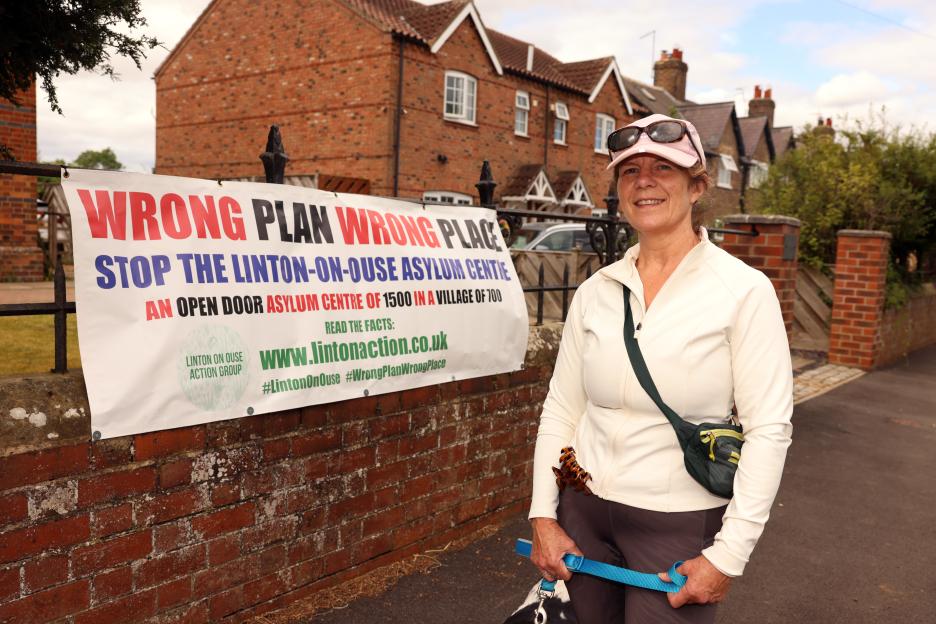 Local resident Della Blood smiling in front of a protest banner that reads "WRONG PLAN WRONG PLACE STOP THE LINTON-ON-OUSE ASYLUM CENTRE AN OPEN DOOR ASYLUM CENTRE OF 1500 IN A VILLAGE OF 700".