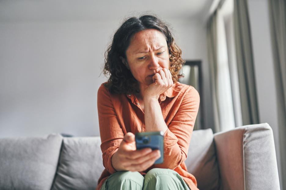 A concerned woman sits on a couch, holding a smartphone and looking at it with a worried expression.