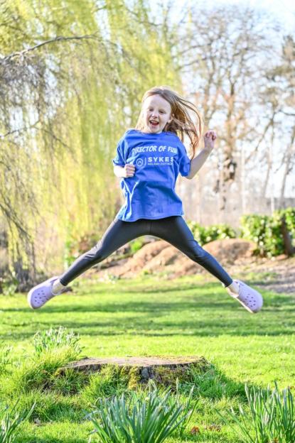 A young girl in a blue shirt with "Director of Fun" written on it jumps in the air.