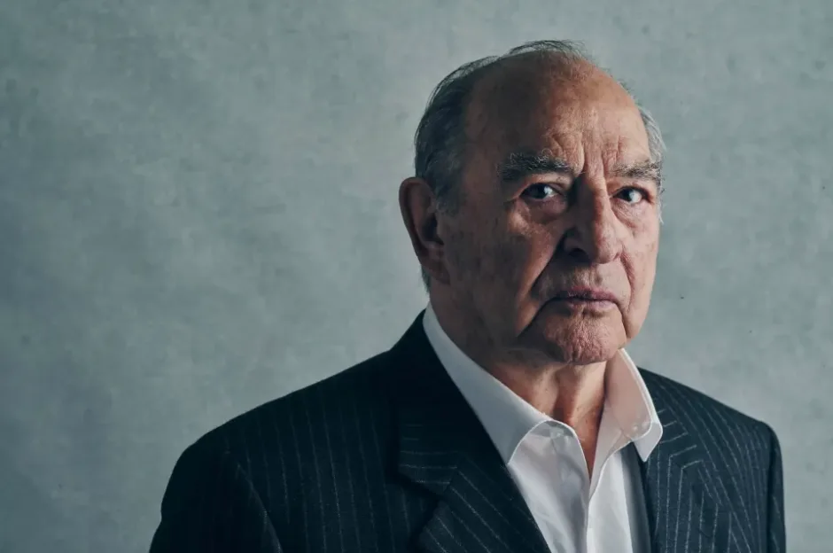 Portrait of a serious older man in a pinstripe suit and white shirt, looking towards the camera against a textured gray background.